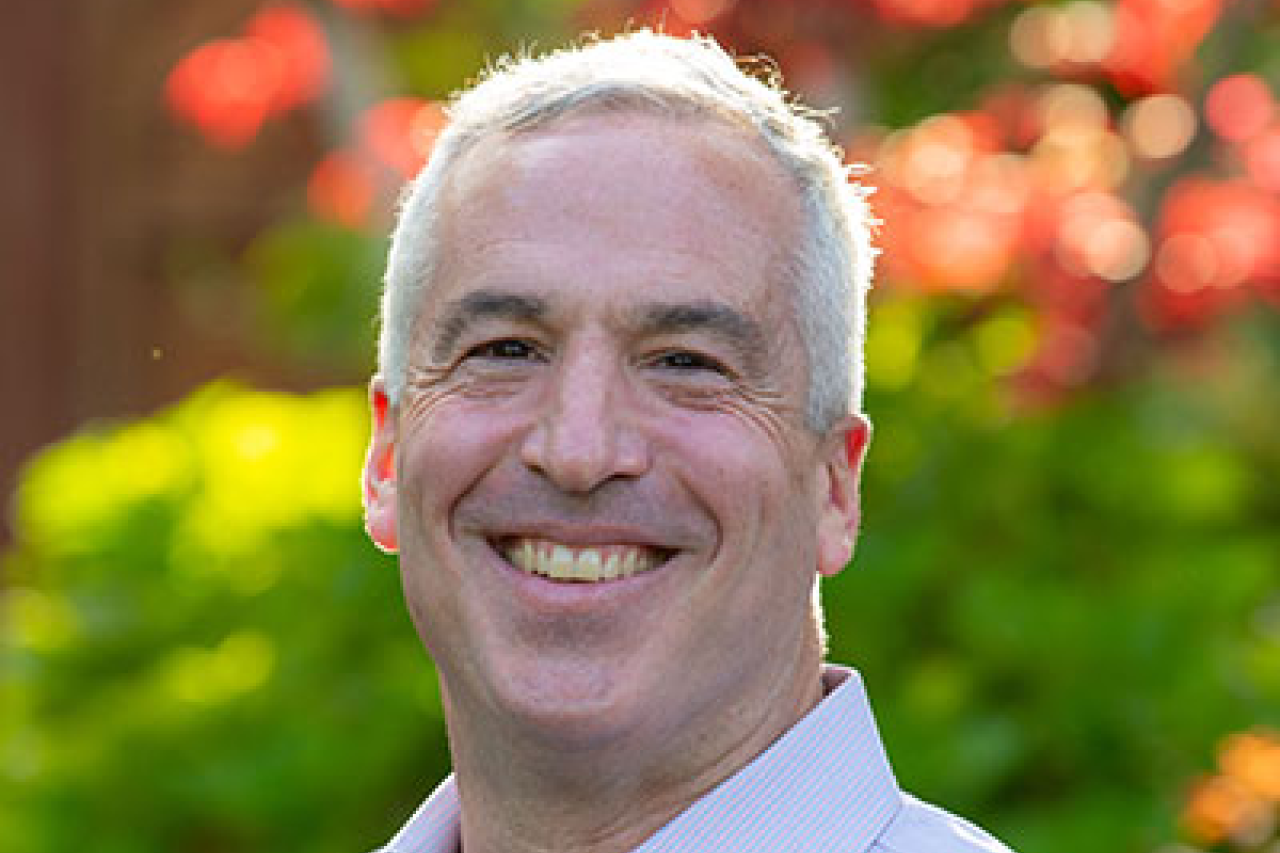 A man in a light blue shirt stands outdoors with lush greenery and blooming flowers.