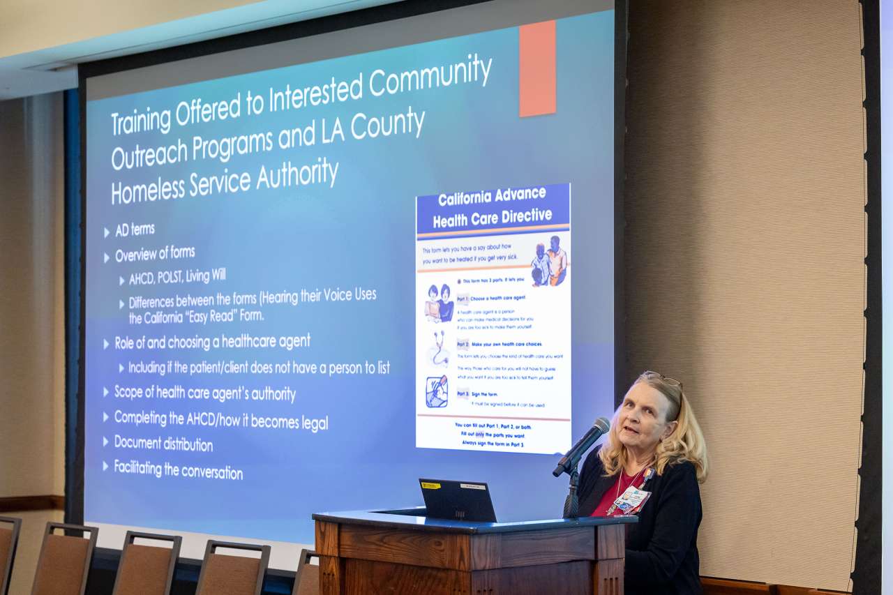 A nurse at a podium speaks at a conference with a Powerpoint slide projected behind her.