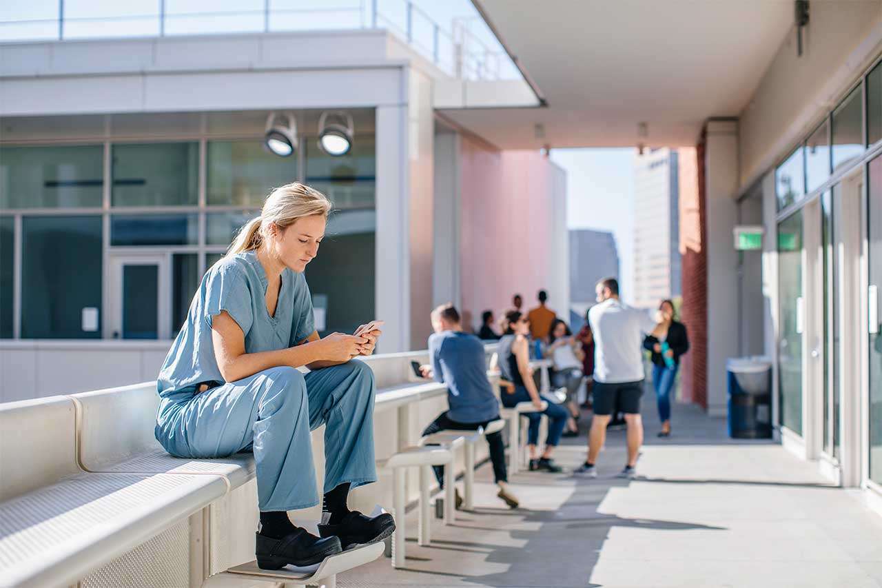 A medical student in scrubs sits on a bench using a smartphone, with people in the background on a terrace.