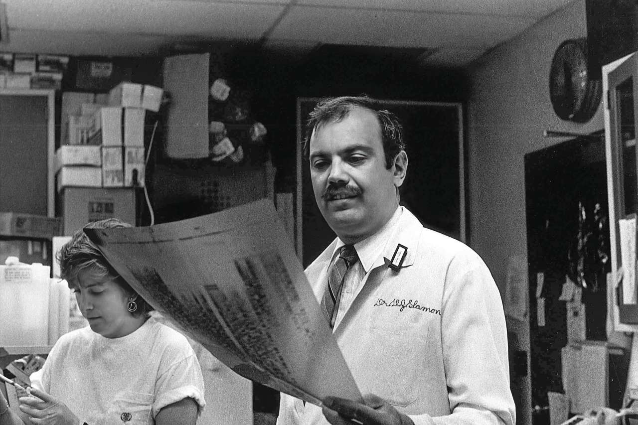 Man in a lab coat labeled "Dr. D.J. Slamon" examines a large printout while a woman beside him works with lab materials.