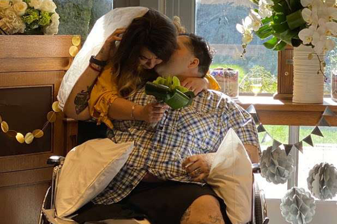 A 3 Wishes patient in a wheelchair embraces his spouse, who is leaning over him, both smiling with eyes closed.