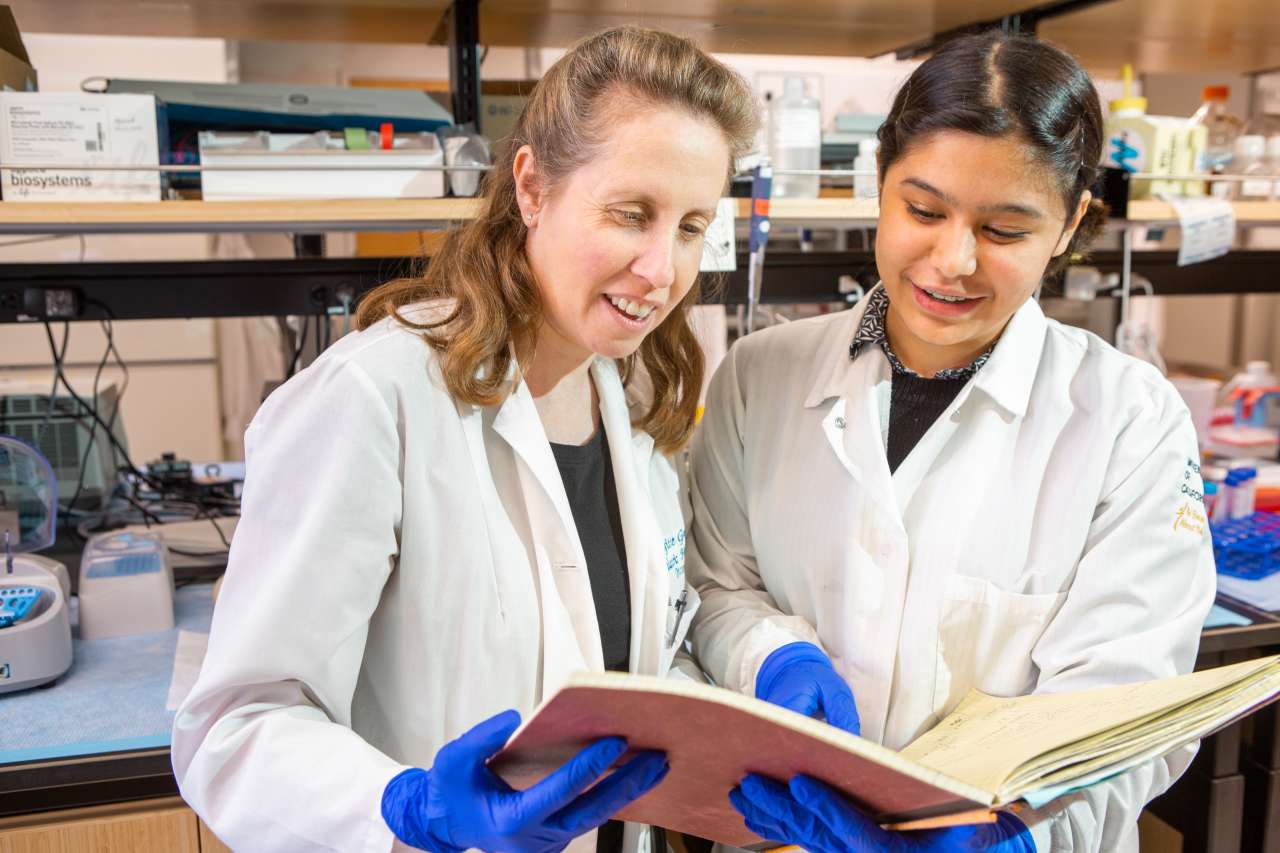 Two smiling women in lab coats and blue gloves look at a book together in a lab