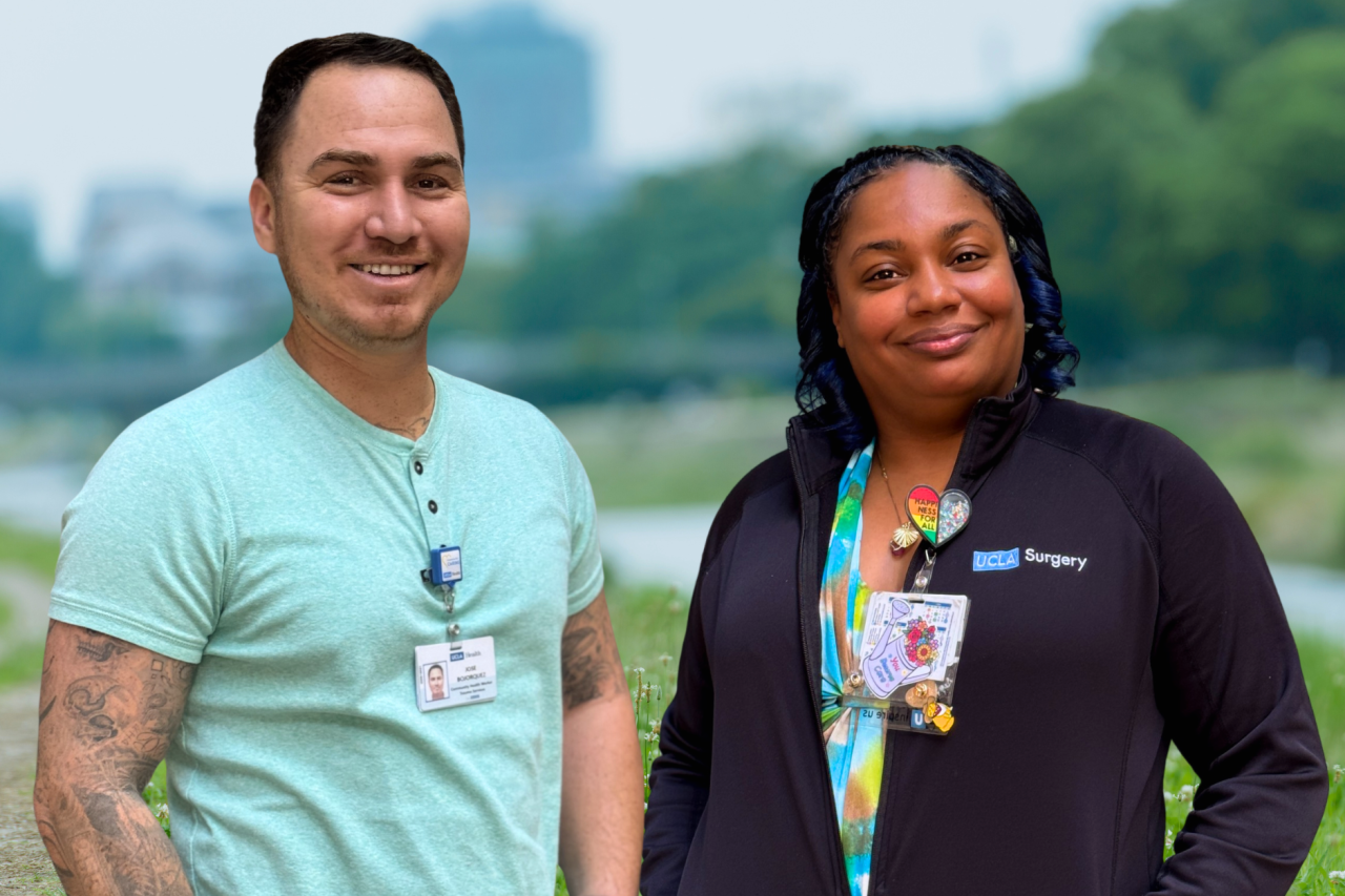 Two UCLA Health employees smiling outdoors, wearing ID badges. One wears a light green shirt, the other a black UCLA Surgery jacket.