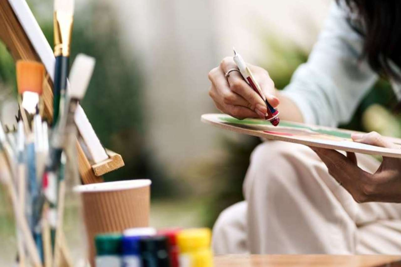 Hands holding a paintbrush and palette, paint jars and brushes on table