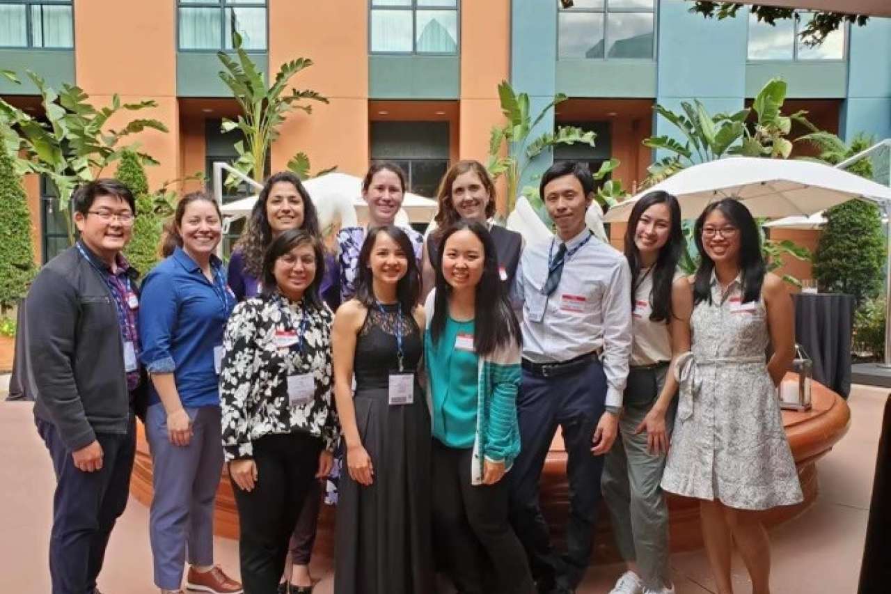 Group of ten people smiling outdoors, standing together in front of a building with plants and umbrellas.