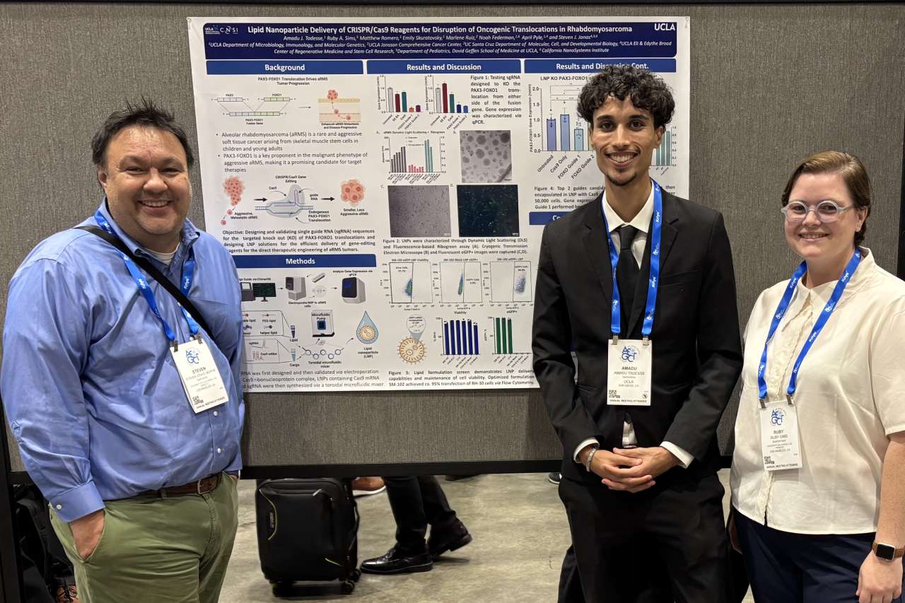 Amadu Tadesse stands with two colleagues in front of a research poster at a scientific conference. All three wear conference lanyards; Amadu is dressed in a suit and tie.