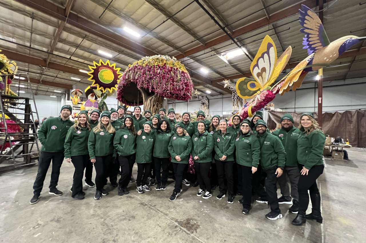 Group of people in green jackets posing with colorful parade floats in a warehouse.
