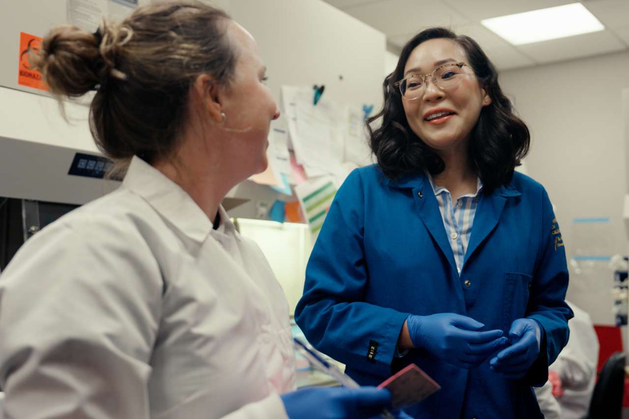 Two researchers in a laboratory smiling and talking, wearing lab coats and gloves.