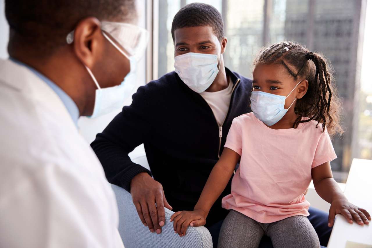 A doctor wearing goggles and mask is facing a young girl and dad with masks.