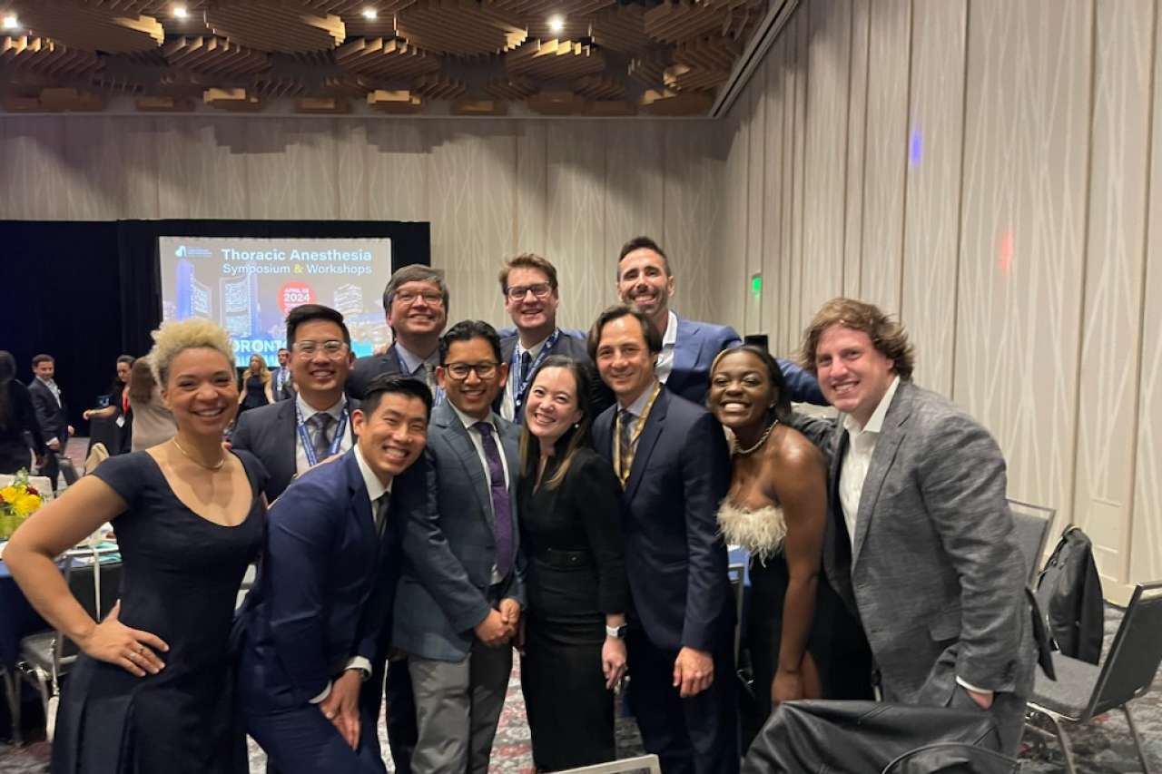 A diverse group of smiling adults, dressed in formal attire, are posing for a photo at an event. A screen in the background displays "Thoracic Anesthesia Symposium & Workshops."