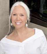 Smiling woman with white hair wearing an off-shoulder white top and dangling earrings.