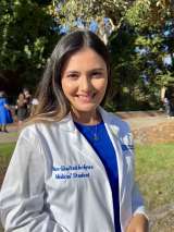 Nare, who has brown hair and is wearing a white coat, blue shirt, and gold necklace posing in front of a tree.