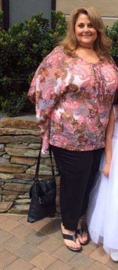 Person wearing a pink patterned blouse, black pants, and sandals standing outdoors beside a child in a white dress.