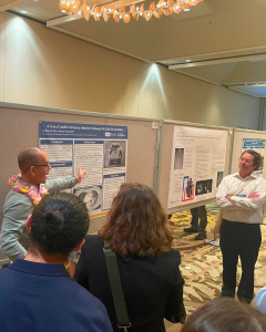 A presenter discusses research next to a poster while attendees listen in a conference setting.