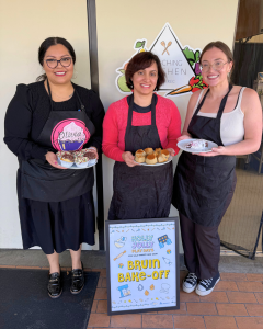Three women holding trays of baked goods, standing outside a kitchen with a sign.