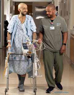 Chaz Valentine accompanies a patient using a walker in a hospital hallway.