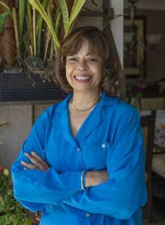 Gail Wyatt a bright blue shirt stands indoors with arms crossed, smiling in front of potted plants.