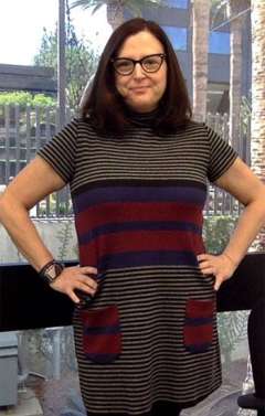 Person standing with hands on hips wearing a striped short‑sleeve tunic with red and blue accents, indoors with large windows behind them.
