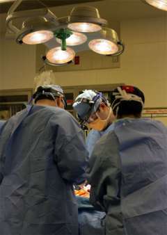  Three surgeons in blue scrubs and head coverings stand around an operating table, illuminated by a large surgical light overhead.