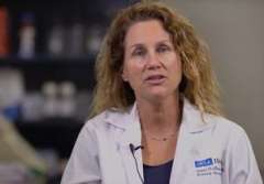 A person in a lab coat speaks in a laboratory setting.