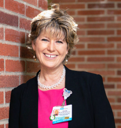 Coleen Wilson in a black blazer and pink top, standing by a brick wall.