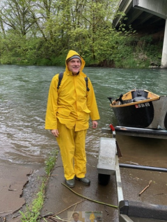 a screenshot of a man next to a river and small boat, with a yellow rainjacket