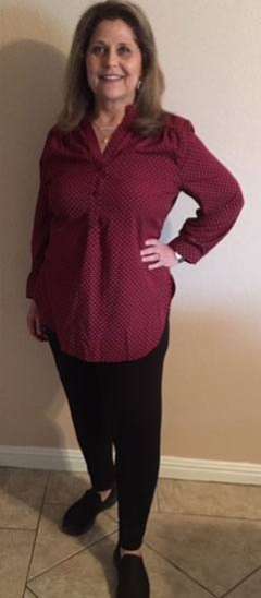 Person standing indoors wearing a long-sleeve red patterned blouse, black pants, and black shoes with one hand on their hip.