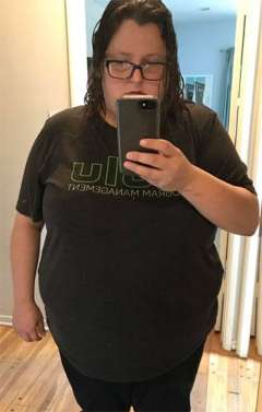 Person taking a mirror selfie wearing a dark T‑shirt and black pants, standing in a hallway with wooden floors.