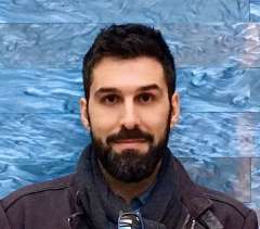 Dr. Jiannis Taxidis, a man with dark hair and a beard, looks forward confidently in a jacket and dark scarf against a blue wall.