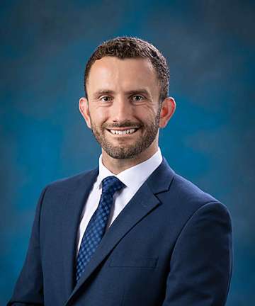 Smiling man in navy suit and tie against blue studio background.