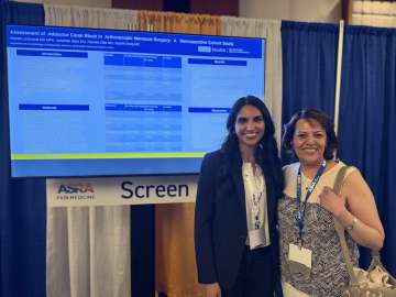 Two people wearing lanyards stand before a large presentation screen and booth curtains.