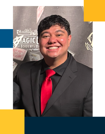 Josh is smiling while standing in front of a magic castle background in a black suit with a red tie