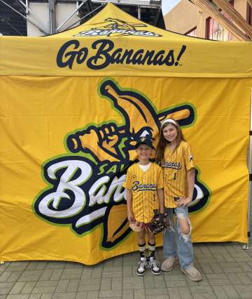 Two people in yellow baseball jerseys standing in front of a yellow "Go Bananas" banner.
