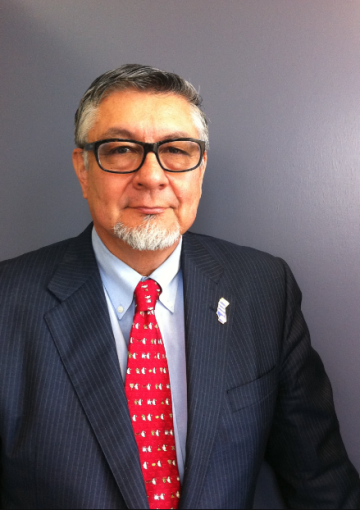 David Hayes-Bautista wearing glasses, a light blue button up shirt, a red tie, and a black suit with thin stripes.