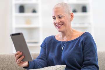 A woman with cancer is undergoing treatment. She has a shaved head because chemotherapy has caused hair loss. The woman is sitting on a couch in her living room at home. She is using a digital tablet to video chat with friends and family. She is smiling joyfully.
