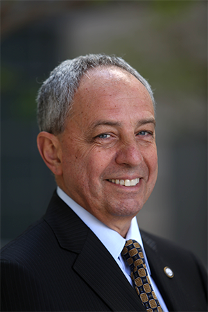 John Mazziotta headshot. Similing to the camera, wearing a black suit, white button up shirt, and gold and black tie.