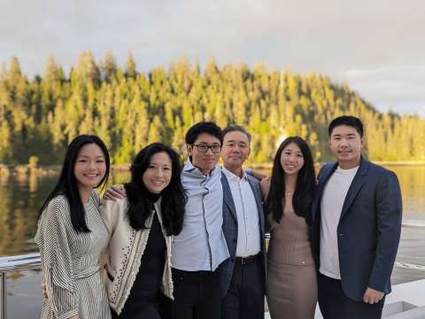 Six adults posed together on a lakeside dock with forested shore behind.