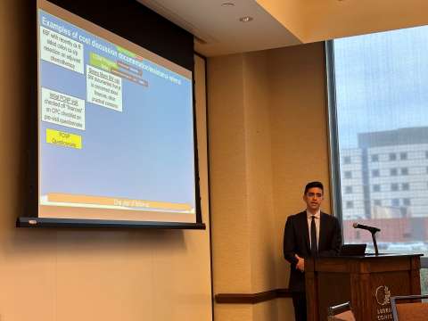 Presenter in suit at podium next to projected slide in a conference room.
