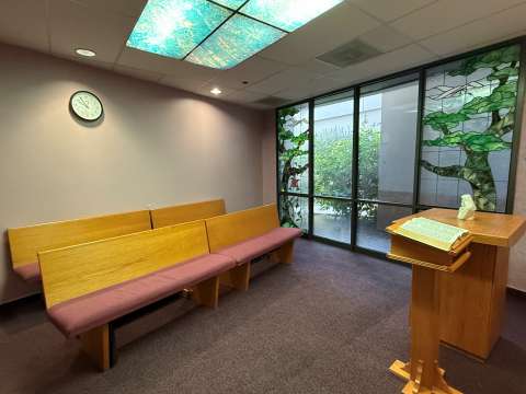 A peaceful chapel room with three wooden pews, a lectern, and a small statue. Stained glass windows with a tree design let in light.