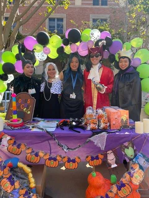 Five individuals dressed in Halloween costumes stand at a festive booth with decorations.