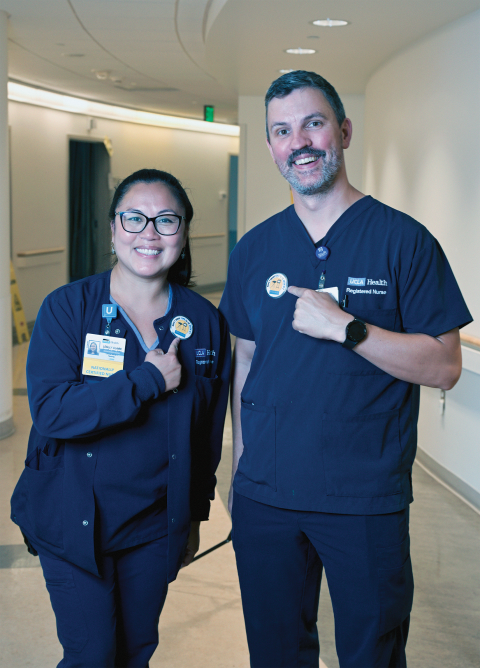 Two nurses in blue scrubs, each wearing a name badge, smiling and standing together.