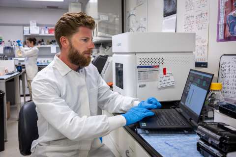 Lab technician working on a laptop in a laboratory setting, wearing gloves and a lab coat.