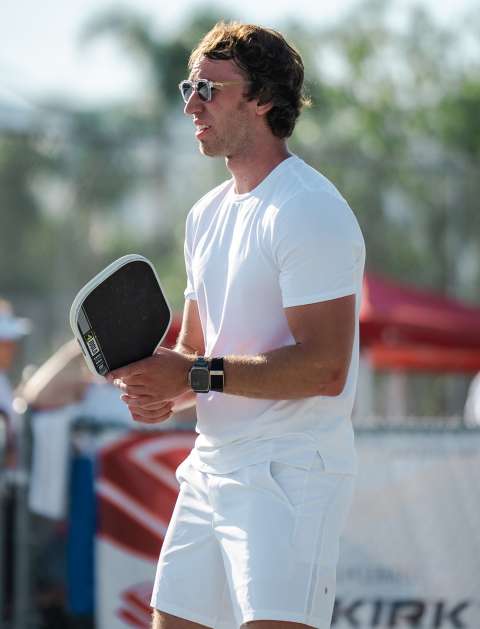 Man in white athletic clothes holding a paddle on an outdoor court.