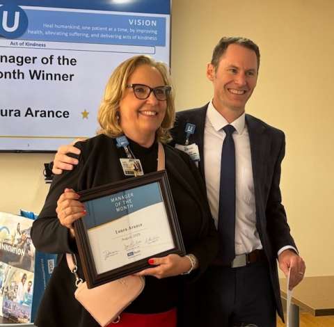  Laura Arance, a smiling woman in glasses, holds a framed award while a man in a suit stands behind her with his hand on her shoulder.