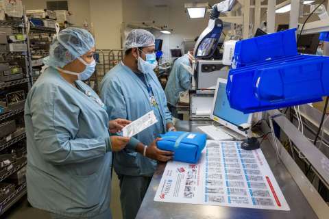 Two healthcare workers in scrubs and hairnets review a checklist while handling a blue sterile pack at a workstation with trays and a computer.