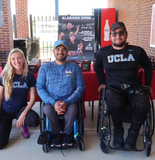  Three UCLA adaptive athletes pose in front of an Alabama Open poster at a sports event venue.