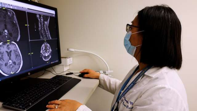Masked clinician examining brain MRI scans on a desktop monitor.