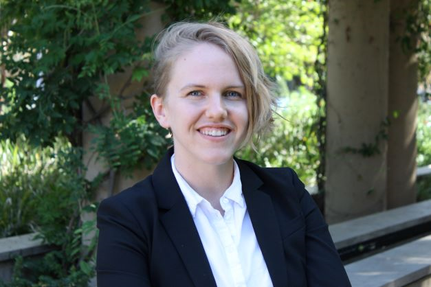 Smiling person with short blonde hair in a black blazer and white shirt, outdoor headshot