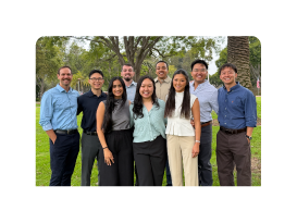 Nine adults standing and smiling outdoors on grass beneath trees.