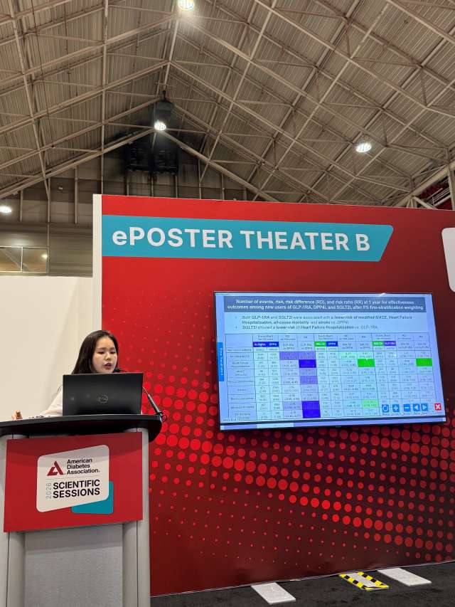 Presenter at ePOSTER Theater B podium beside large screen displaying poster schedule